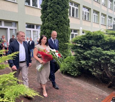 delegacja trzech osób-dwóch mężćzyzn i kobieta która trzyma kwiaty idą pod pomnik