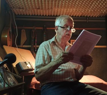 mężczyzna siedzi na stołku w malym studio nagraniowym, trzyma kartki w dłoniach, czyta tekst