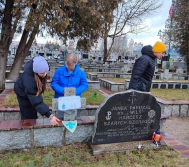 Na dworze, cmentarz. Przy grobie dwie nastolatki dekorują krzyż. Nastolatek w czarnej kurtce i żółtej czapce odchodzi