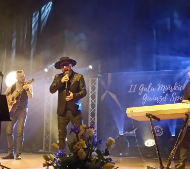 Na scenie mężczyzna w kapeluszu i czarnych okularach śpiewa do mikrofonu. Po lewej na gitarze gra mężczyzna. W tle duży...
