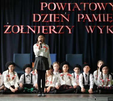 Aula szkolna. Na scenie grupa dzieci siedzi po turecku, na przodzie dziewczynka. Wszyscy w czarnych beretach, w bialych...