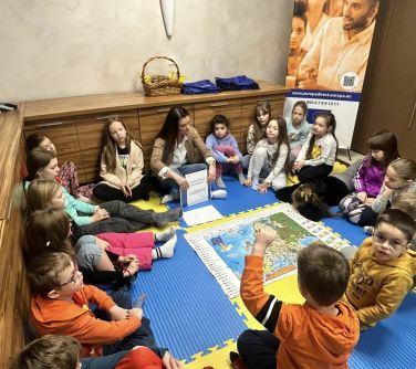 Pomieszcznie. Na kolorowych, piankowych matach kobieta z dziećmi siedzą po turecku. na środku duża mapa. Kobieta opowiada