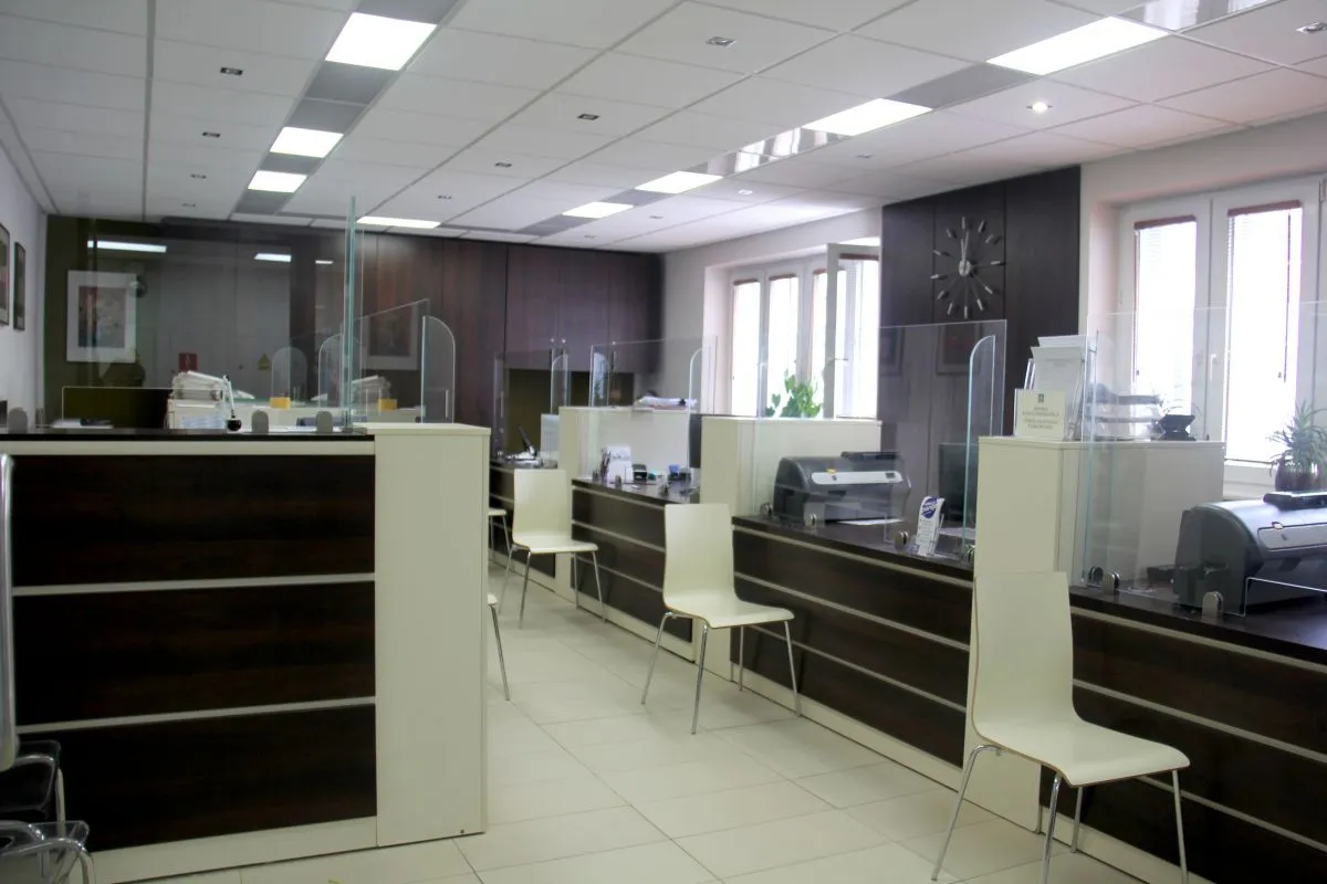 Kliknięcie powiększy obraz. Recepcja z białymi krzesłami, przegrodami ochronnymi z przezroczystego szkła oraz ciemnymi meblami i sprzętem biurowym.