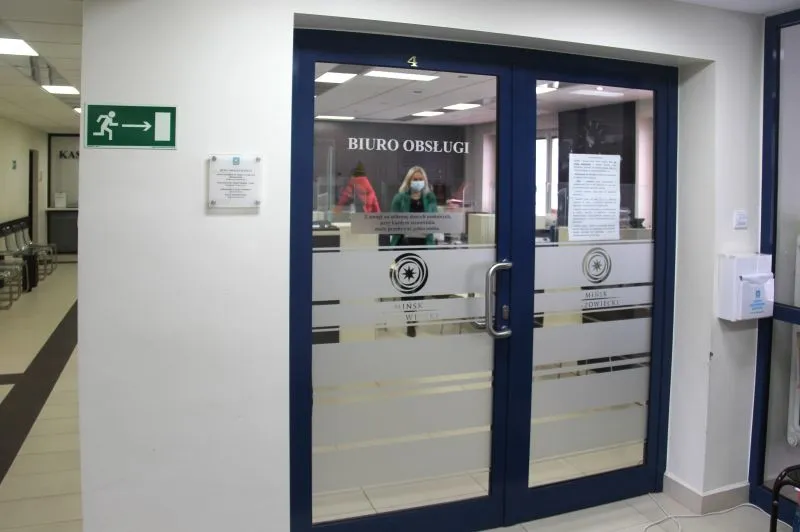 Kliknięcie powiększy obraz. Dwuskrzydłowe drzwi z napisem "BIURO OBSŁUGI" oraz osoba nosząca maseczkę stojąca za szybą.