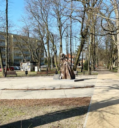 Rzeźba kobiety w sukni na postumencie w parku z ławkami i drzewami bez liści.