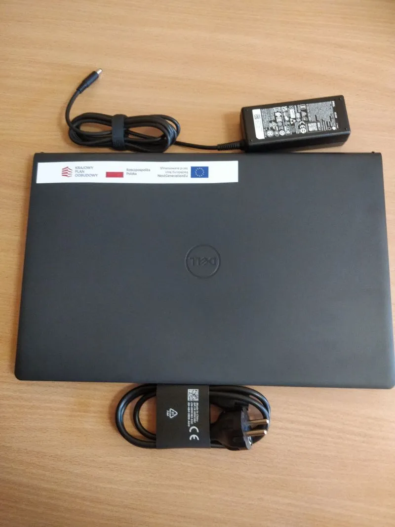 Czarny laptop marki Dell z naklejką programu Krajowy Plan Odbudowy, zasilacz i przewód zasilający na drewnianym blacie.
