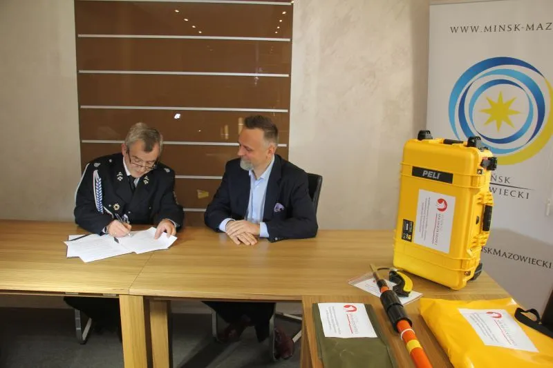 Dwóch mężczyzn siedzi przy stole, jeden podpisuje dokumenty, obok stoi żółta walizka na sprzęt i leży sprzęt ratowniczy.