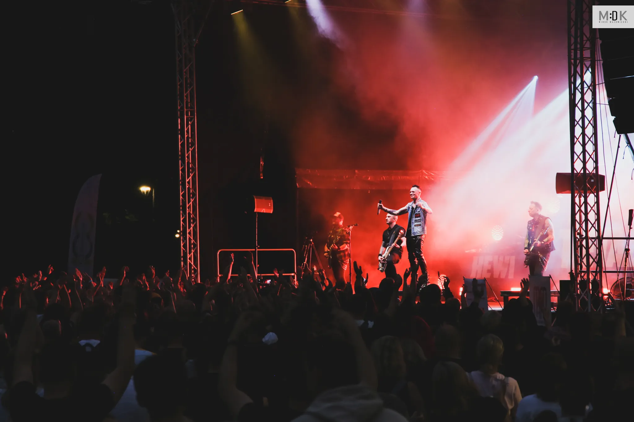 Zespół muzyczny gra na scenie oświetlonej czerwonym światłem, a przed nimi stoi tłum widzów z uniesionymi rękami.