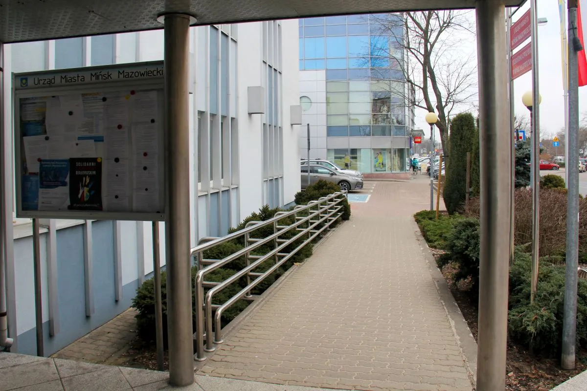 Tablica informacyjna Urzędu Miasta Mińsk Mazowiecki obok budynku z chodnikiem i barierkami.