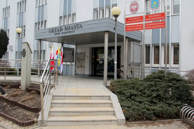 Wejście do budynku Urzędu Miasta Mińsk Mazowiecki z podjazdem, schodami oraz tablicami informacyjnymi i flagami.