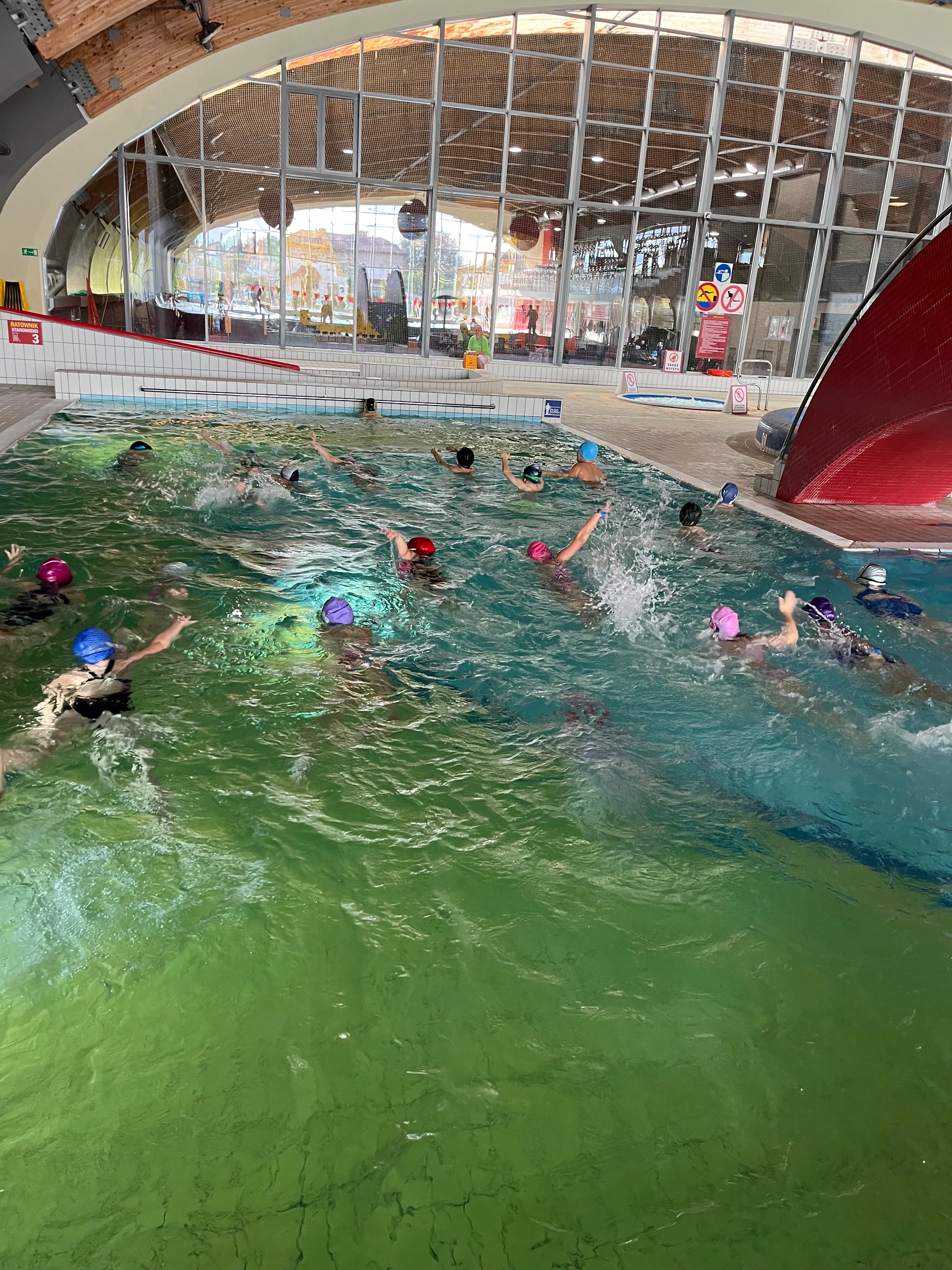 Wewnętrzny basen pływacki z grupą dzieci pływających w kolorowych czepek. W tle szklana ściana z widokiem na halę rekreacyjną i drewnianym sklepieniem. Obok basenu czerwony zjeżdżalnia wodna i osoba siedząca przy krawędzi basenu.
