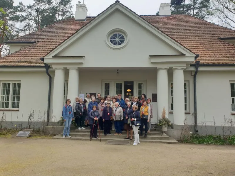 Grupa osób stoi na schodach przed budynkiem z białymi ścianami, dwiema kolumnami i okrągłym oknem w szczycie dachu.