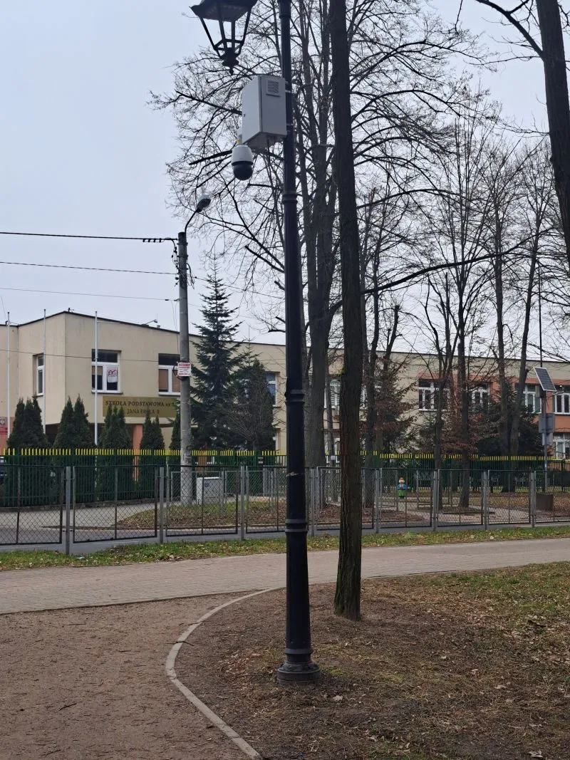Latarnia uliczna z zamontowaną kamerą monitoringu i drzewami wokół na tle budynku szkoły.
