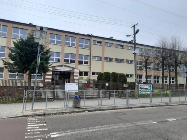 Trzypiętrowy budynek szkolny z licznymi oknami i ogrodzeniem przed wejściem od ulicy.