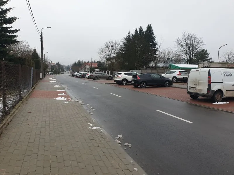 Ulica z chodnikiem i zaparkowanymi samochodami po prawej stronie oraz rozproszonymi resztkami śniegu na chodniku i ulicy.