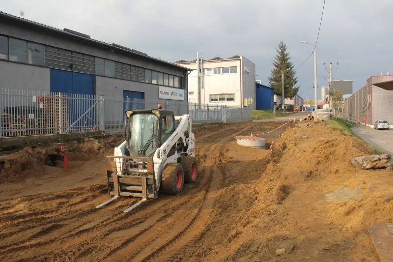 Maszyna budowlana na terenie wykopu wzdłuż zabudowań przemysłowych.