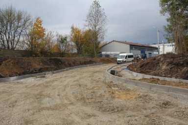 Budowa drogi z widocznym korytem i krawężnikami, biała furgonetka oraz budynki w tle.