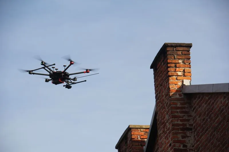 Czarny wielowirnikowy dron latający obok czerwonego ceglastego komina na tle jasnego nieba.