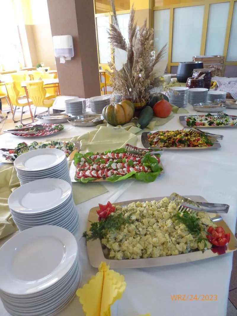 Stoły z nakryciami, kilkoma talerzami sałatek oraz dekoracją z traw i warzyw.