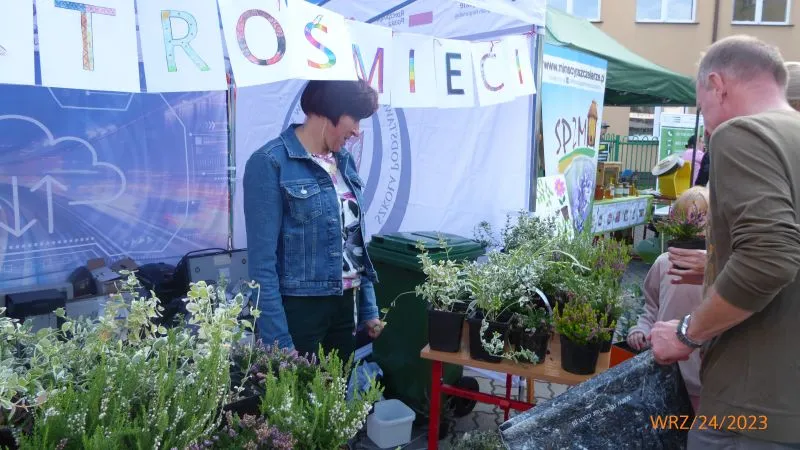 Stoisko z roślinami doniczkowymi i dwie osoby rozmawiające przy tym stoisku pod banerem z napisem "Trosmiec".