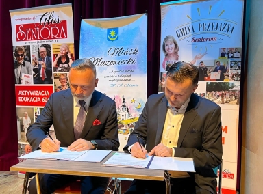 Dwaj mężczyźni w garniturach podpisują dokumenty siedząc przy stole, za nimi stoją banery promujące inicjatywy dla seniorów.