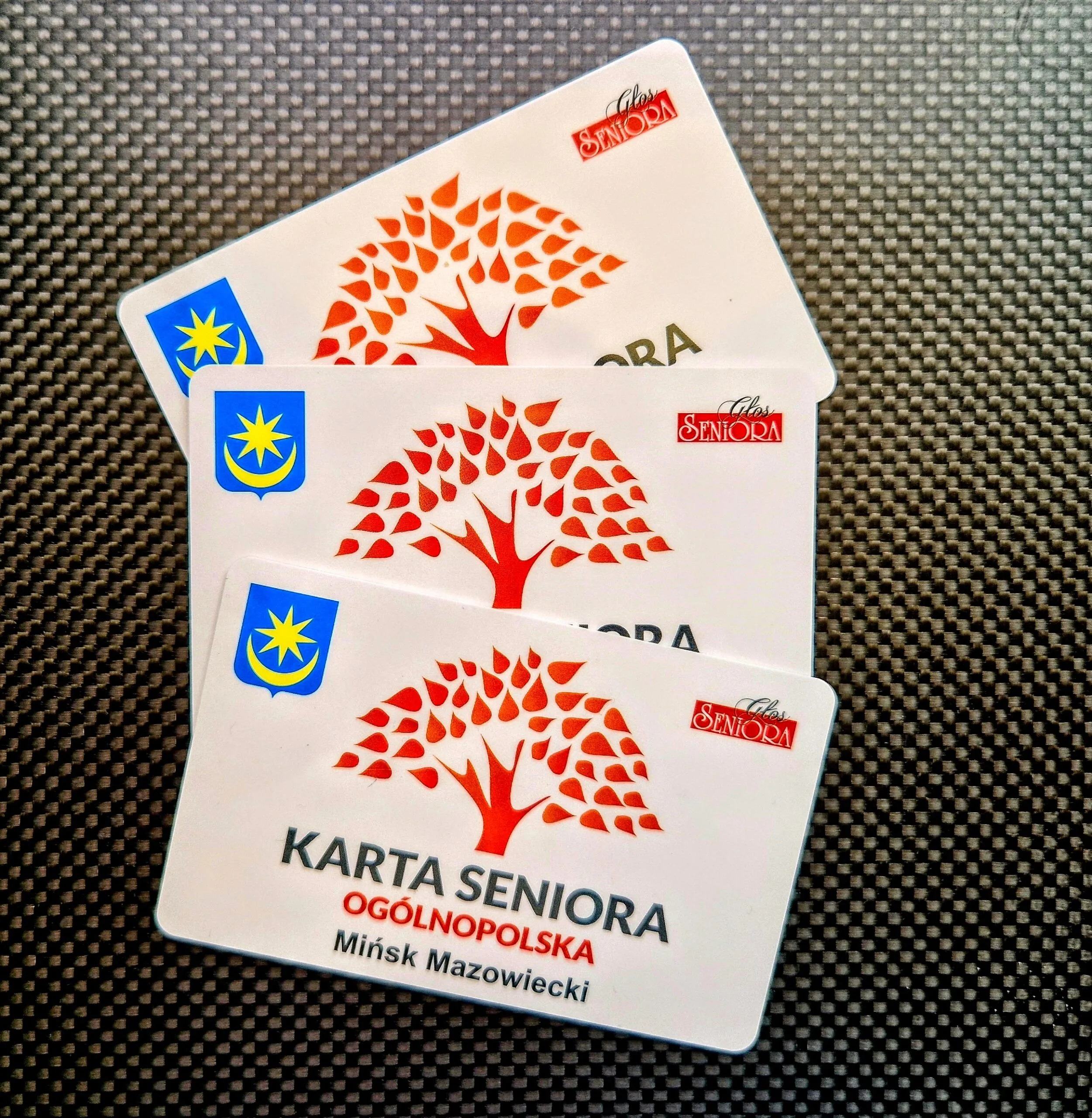 Trzy białe karty „Karta Seniora Ogólnopolska” z czerwonym logo drzewa i symbolem herbu Mińska Mazowieckiego na czarnym tle.
