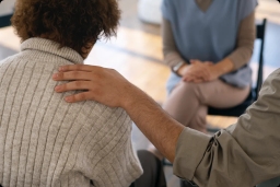 Ręka osoby dotykająca ramię drugiej osoby w szarym swetrze, z trzecią osobą siedzącą naprzeciwko.