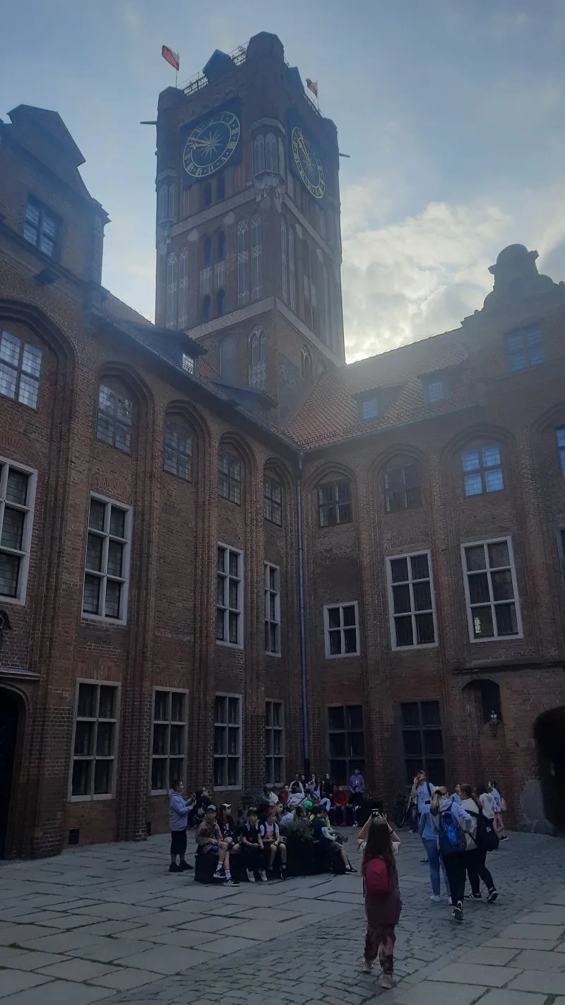 Zabytkowy ceglany budynek z wysoką wieżą zegarową oraz grupa turystów w dziedzińcu.