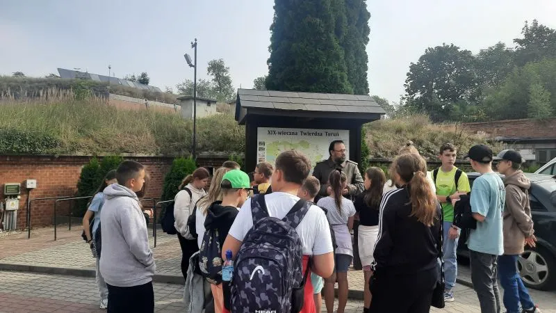 Grupa dzieci i młodzieży z plecakami stoi przed tablicą informacyjną pod zadaszeniem na zewnątrz.