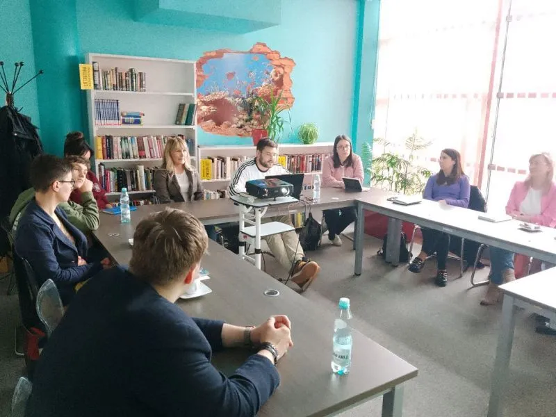 Grupa osób siedzących przy stołach ułożonych w kształt litery U w sali z regałami z książkami i projektorem.