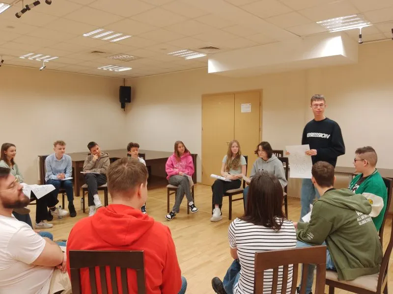 Grupa młodych ludzi siedzi na krzesłach w kółku, a jedna osoba stoi i prezentuje kartkę z notatkami.