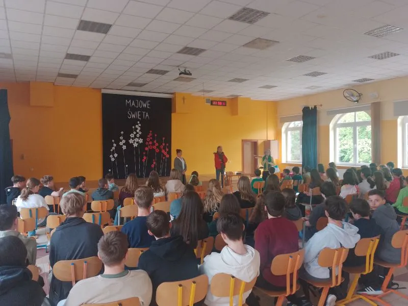 W sali z żółtymi ścianami siedzi grupa młodzieży zwrócona w stronę trzech osób stojących przed czarną dekoracją z napisem "Majowe święta".