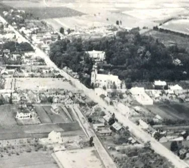 Kliknięcie powiększy obraz. Widok z lotu ptaka na wiejskie zabudowania z drogą przebiegającą przez centrum oraz kościołem otoczonym drzewami.