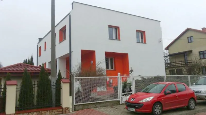 Dwupiętrowy biały budynek z pomarańczowymi akcentami, ogrodzony siatką, z czerwonym samochodem zaparkowanym przed bramą.