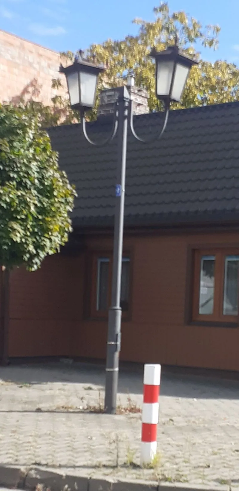 Latarnia uliczna z dwiema lampami na tle domu z ciemnym dachem, obok zielone drzewo i biało-czerwony słupek na chodniku.