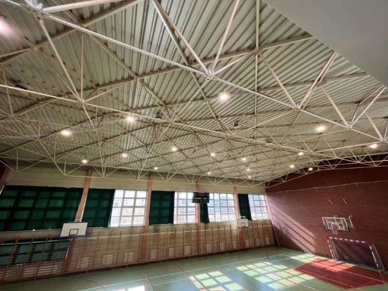 Hala sportowa z boiskiem, trzema tablicami do koszykówki, bramką do piłki ręcznej oraz dużymi oknami na ścianie.