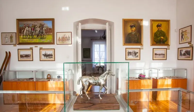 Srebrna figurka konia w szklanej gablocie na tle ściany z obrazami i portretami.
