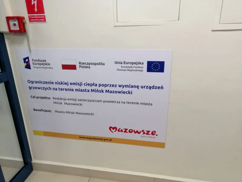 Tablica informacyjna dotycząca projektu ograniczenia emisji ciepła przez wymianę urządzeń grzewczych w mieście Mińsk Mazowiecki z logo Funduszy Europejskich, Polski i Unii Europejskiej.