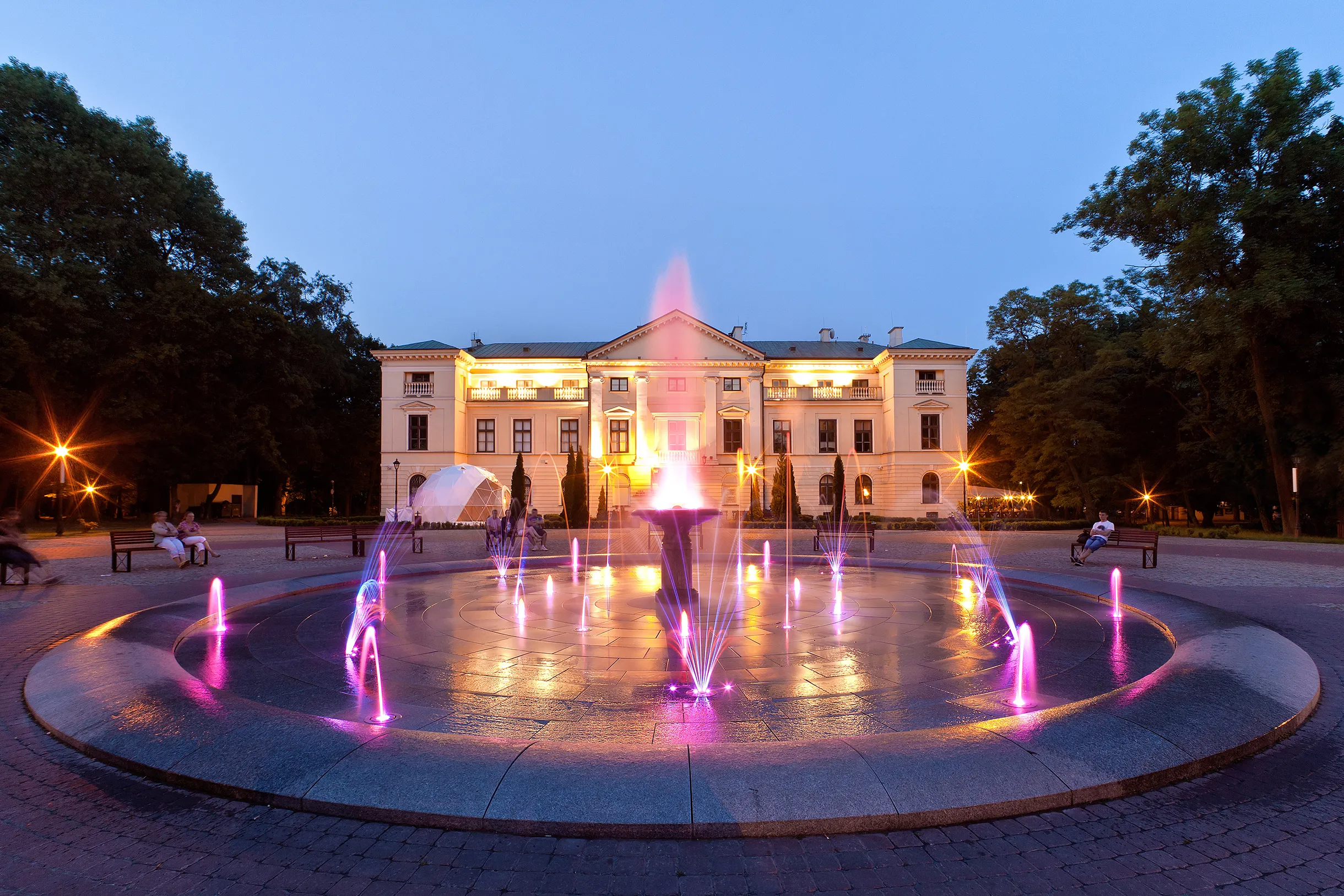 Wieczorny plac z okrągłą fontanną migającą fioletowo-różowym światłem. Woda tryska z kilku dysz i centralnego zbiornika. W tle oświetlony neoklasycystyczny budynek z kolumnami. Po bokach duże drzewa i ławki z kilkoma siedzącymi osobami. Jasne niebo zmierzchu.