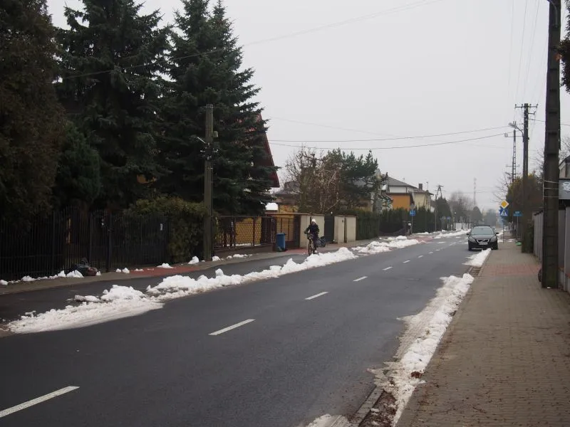 Pusty asfaltowy odcinek ulicy z resztkami śniegu na poboczach, rowerzysta na chodniku i zaparkowany samochód.