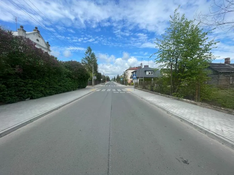 Pusta droga z chodnikami po obu stronach, znak przejścia dla pieszych i zabudowania mieszkalne wzdłuż ulicy.