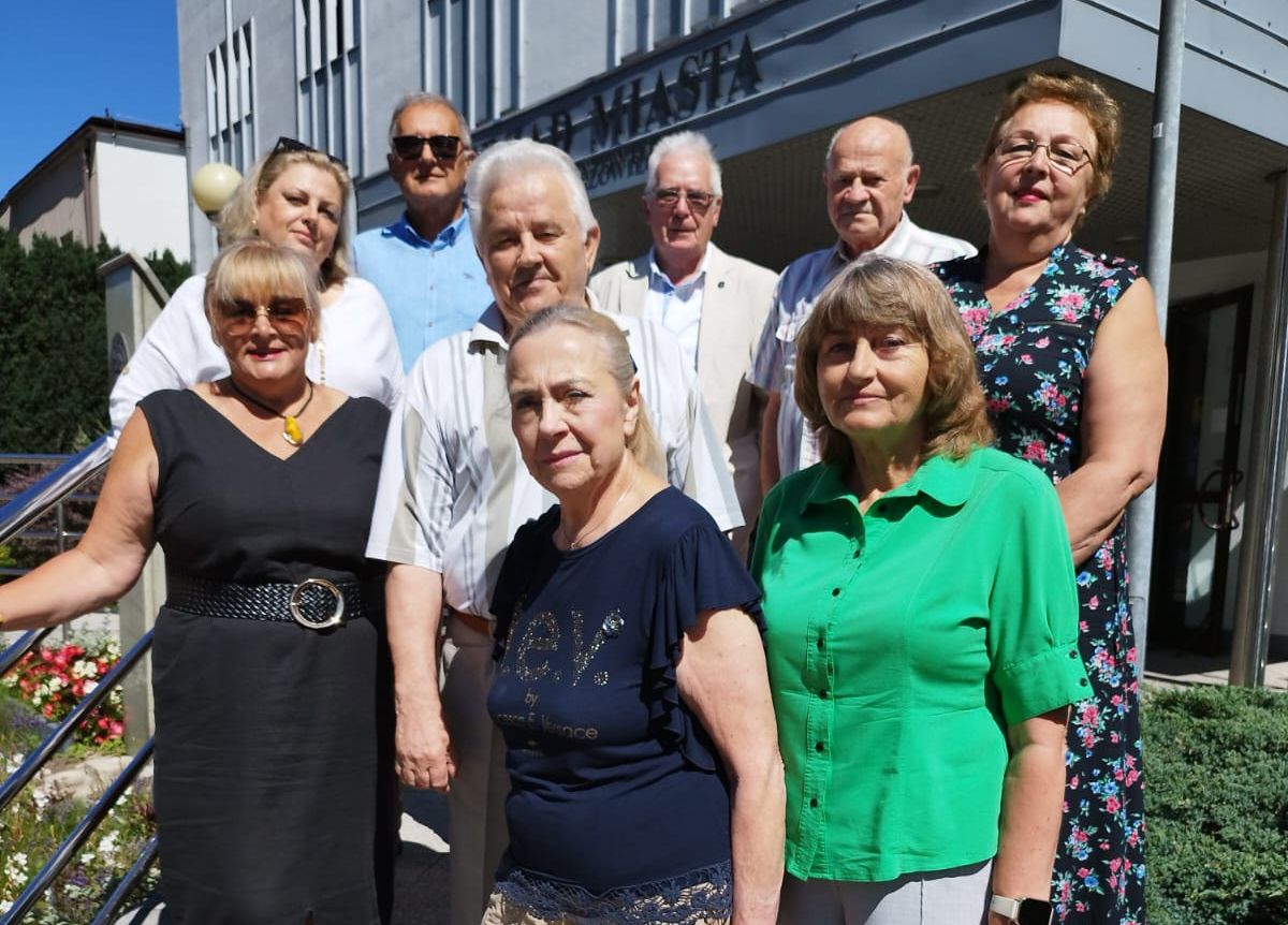 Kliknięcie powiększy obraz. Na zdjęciu grupa dziesięciu osób stoi na zewnątrz, na tle budynku z napisem, w słoneczny dzień. Pięć osób znajduje się z przodu, a pięć z tyłu, w różnych kolorach ubrań.