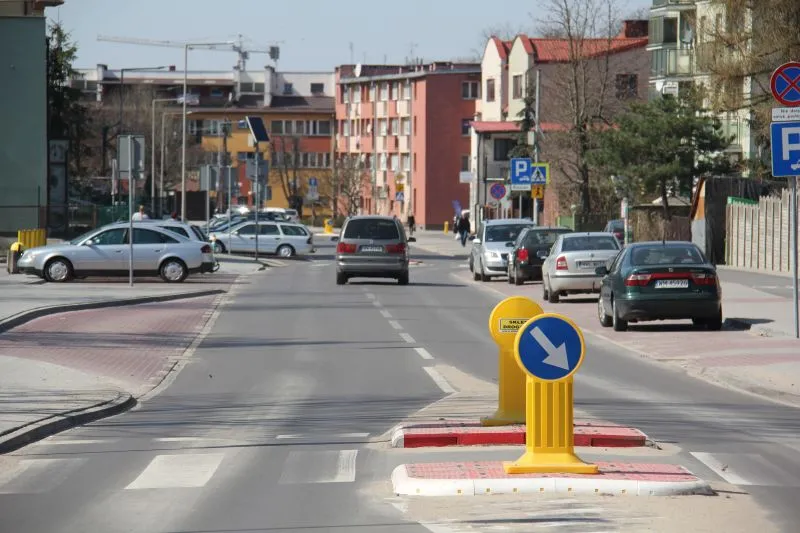 Ulica z pasami dla pieszych, wysepką wymuszającą jazdę po prawej stronie oraz zaparkowanymi samochodami i budynkami mieszkalnymi w tle.