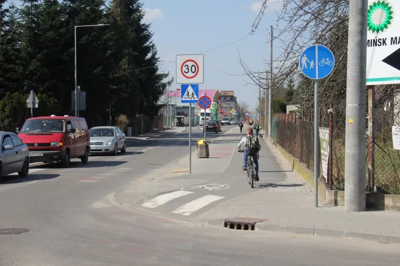 Ulica z ruchem samochodowym po lewej i ścieżką rowerowo-pieszą, na której jedzie rowerzysta, oraz kilkoma znakami drogowymi.