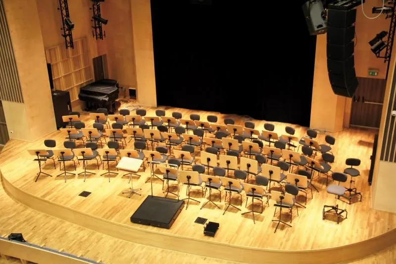 Kliknięcie powiększy obraz. Scena z ustawionymi krzesłami i pulpitami dla muzyków, statywem na nuty oraz głośnikami.