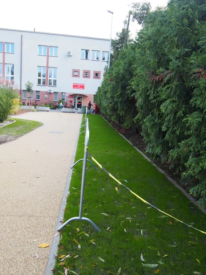 Budynek szkolny z wejściem oznaczonym czerwoną tablicą, obok chodnik i ogrodzony trawnik z żywopłotem.