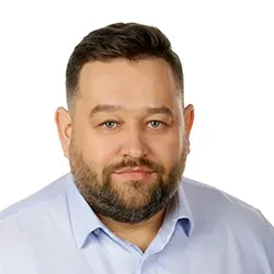 Mężczyzna z krótkimi ciemnymi włosami i brodą ubrany w jasnoniebieską koszulę na białym tle.