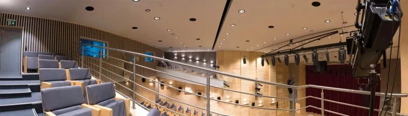 Wnętrze sali teatralnej z widownią, balkonem, sceną zasłoniętą bordowymi kurtynami oraz oświetleniem sufitowym i scenicznym.