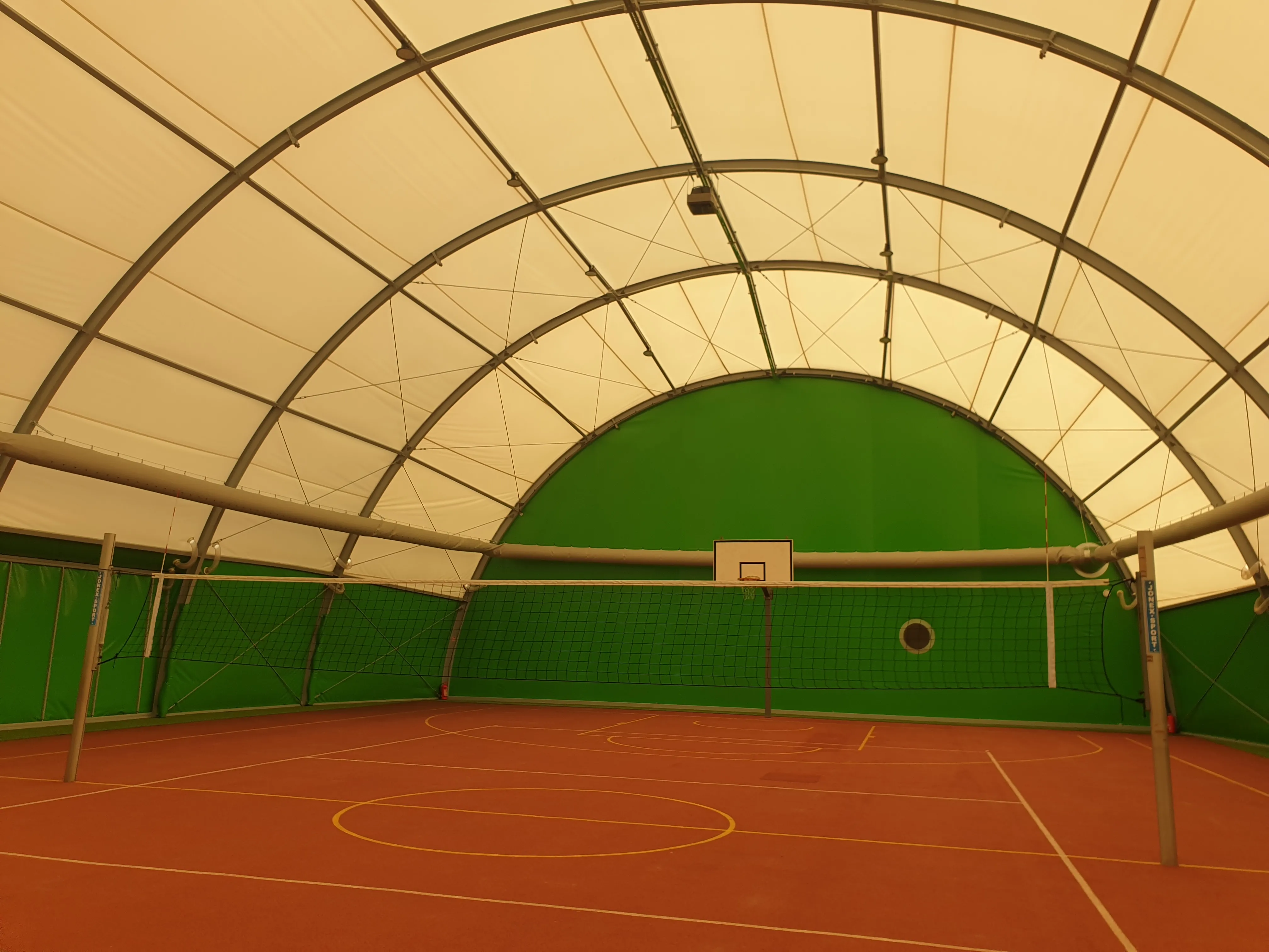 Hala sportowa pod białym, łukowym dachem z metalowymi żeberkami. Zielone ściany i czerwony kort z żółtymi liniami. Siatka do siatkówki rozciągnięta przez środek boiska, kosz zawieszony na tylnej ścianie.