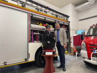 Dwaj mężczyźni stoją przed otwartym wozem strażackim w garażu. Jeden w mundurze strażaka, drugi w marynarce i koszuli. Przed nimi stoi na stoliku duży wentylator. W tle czerwony wóz strażacki.